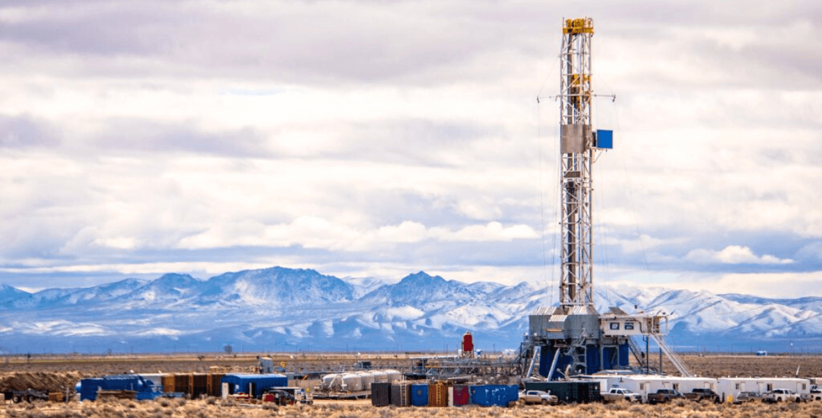 Tall industrial drilling rig in a desert landscape with snow-capped mountains in the background.