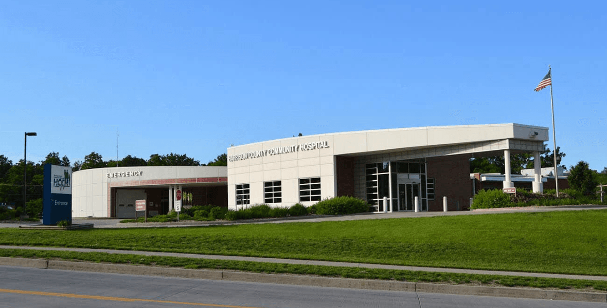 Modern Harrison County Community Hospital exterior with emergency entrance, green lawn, and American flag.