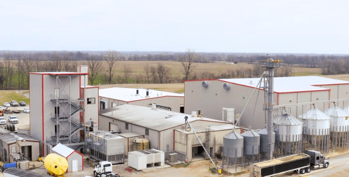 Industrial grain facility with metal buildings, silver silos, and a semi-truck in rural fields.